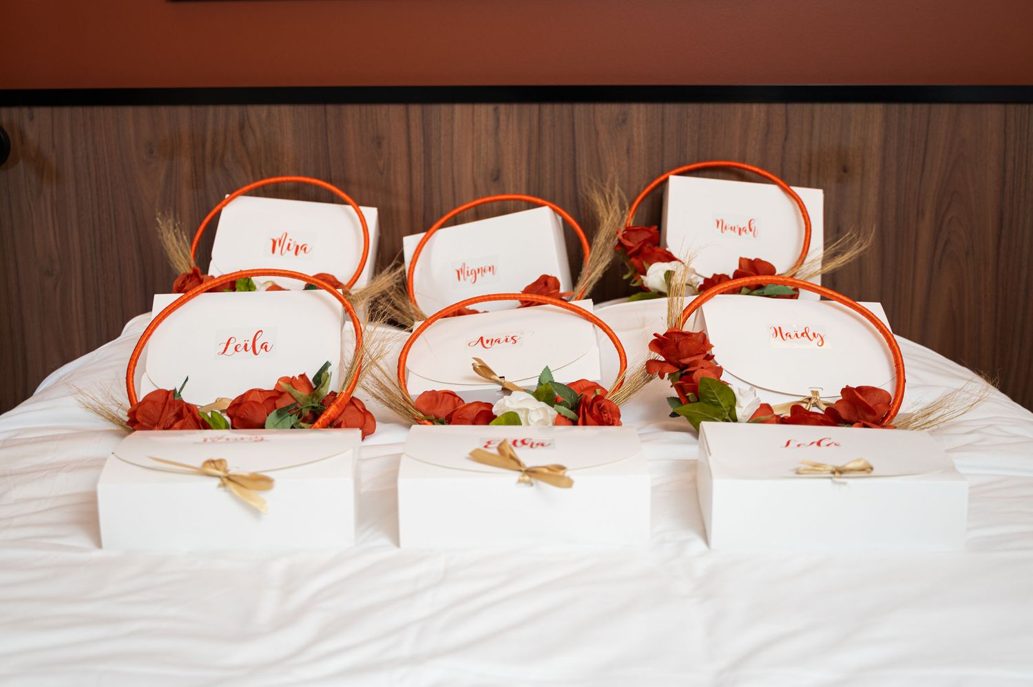 Six personalized white bridesmaid gift boxes with red roses and wheat accents arranged on white bed.
