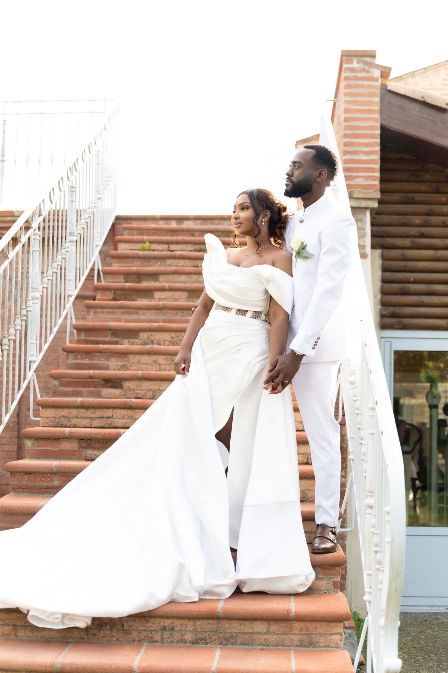 Stunning bride and groom in white wedding attire posing on brick stairs with flowing cathedral train.