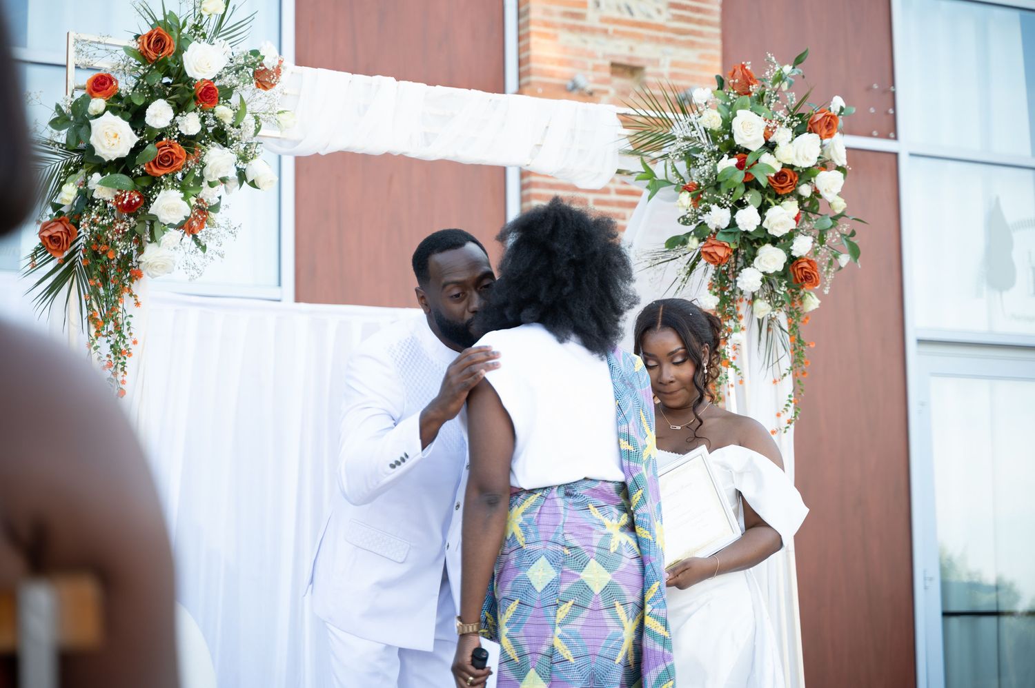 Groom in white suit greets female officiant during outdoor wedding ceremony with bride.