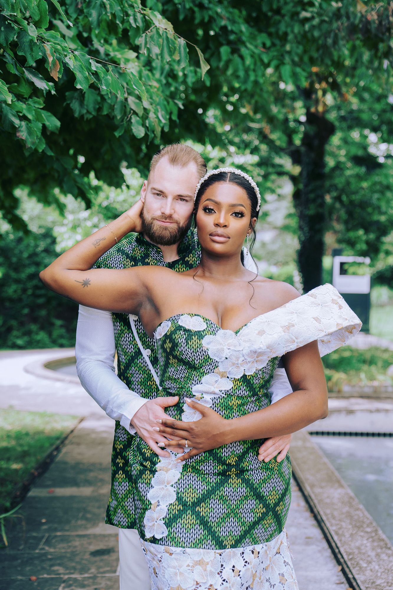 Interracial wedding couple embraces outdoors wearing matching green African print attire among trees.