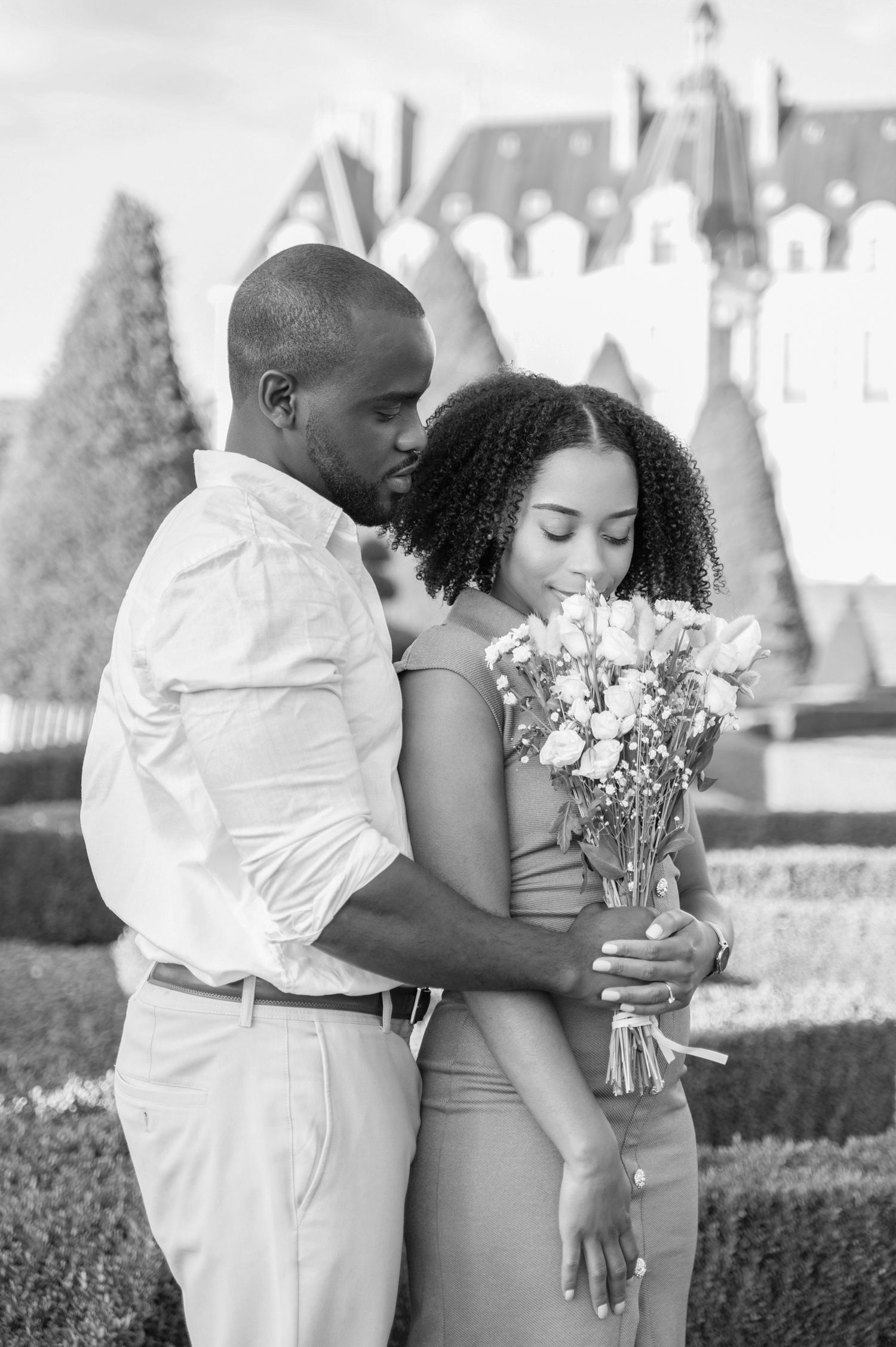 Black and white photo of couple embracing with flower bouquet near formal garden.
