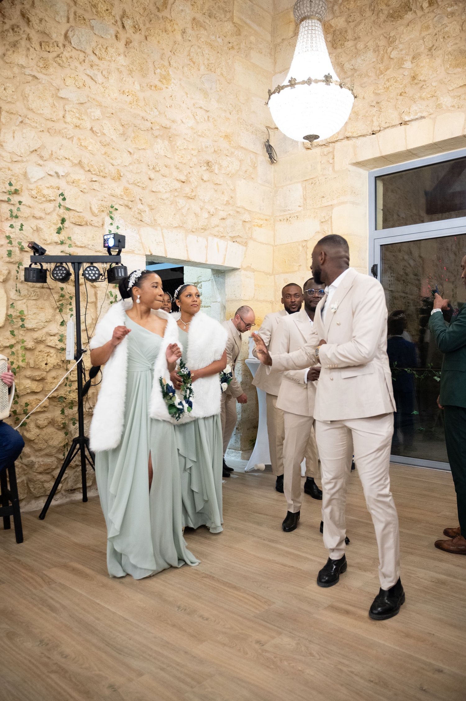A Black groom in a cream suit dances with a bridesmaid in sage green at a stone-walled wedding reception.