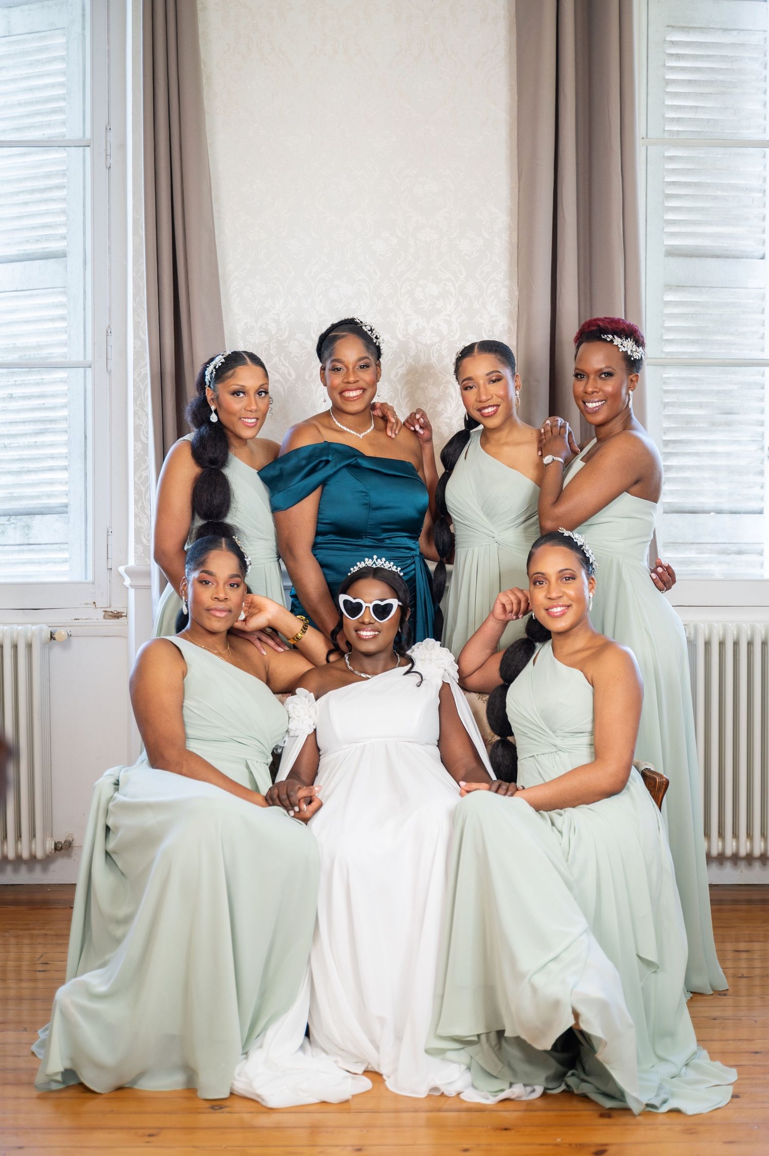 Bride in white dress with sunglasses poses with six bridesmaids in sage green gowns at wedding.