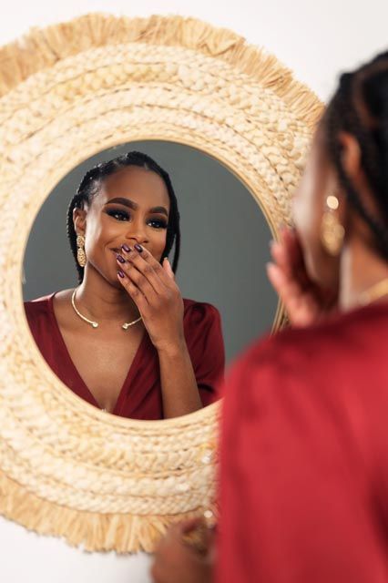 A young Black woman in a red dress admires herself in a round woven mirror, smiling with her hand near her face.