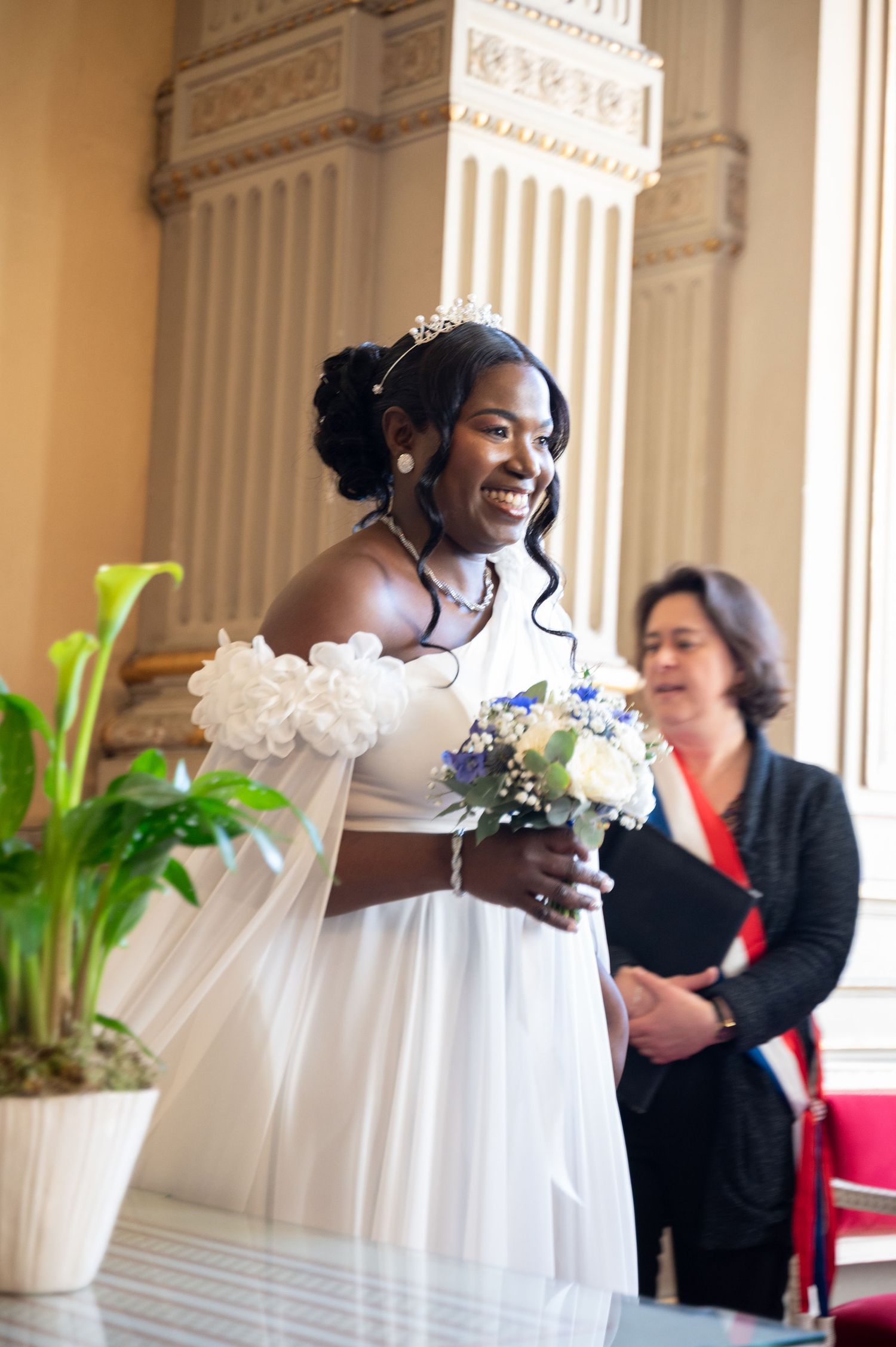 Radiant bride in white off-shoulder gown holding bouquet smiling during civil wedding ceremony.