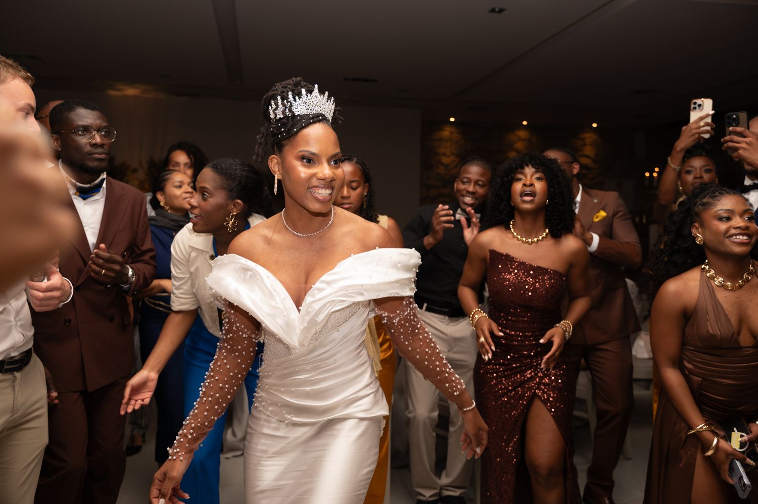 Radiant bride in pearl-sleeve gown and crystal crown celebrates joyfully with guests at wedding reception.