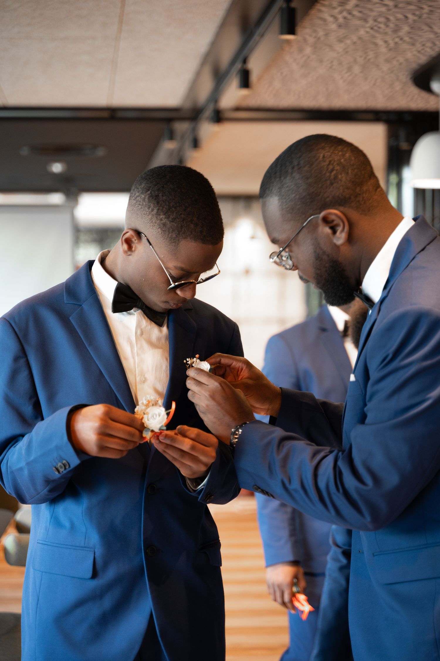 Two Black groomsmen in blue suits carefully pin a boutonniere onto a lapel before a wedding.
