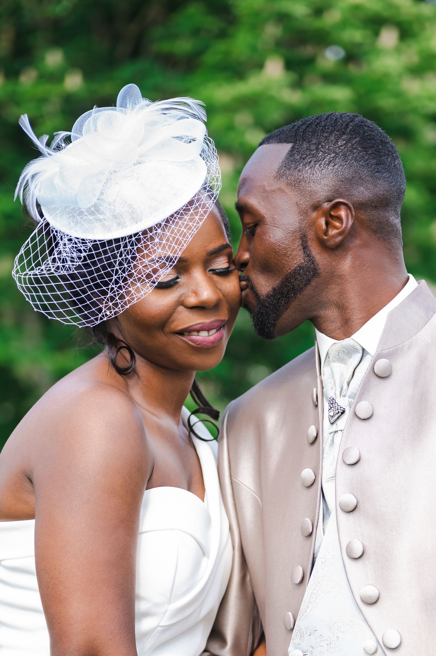 Groom in beige suit kisses bride wearing white fascinator hat and strapless gown in romantic close-up.