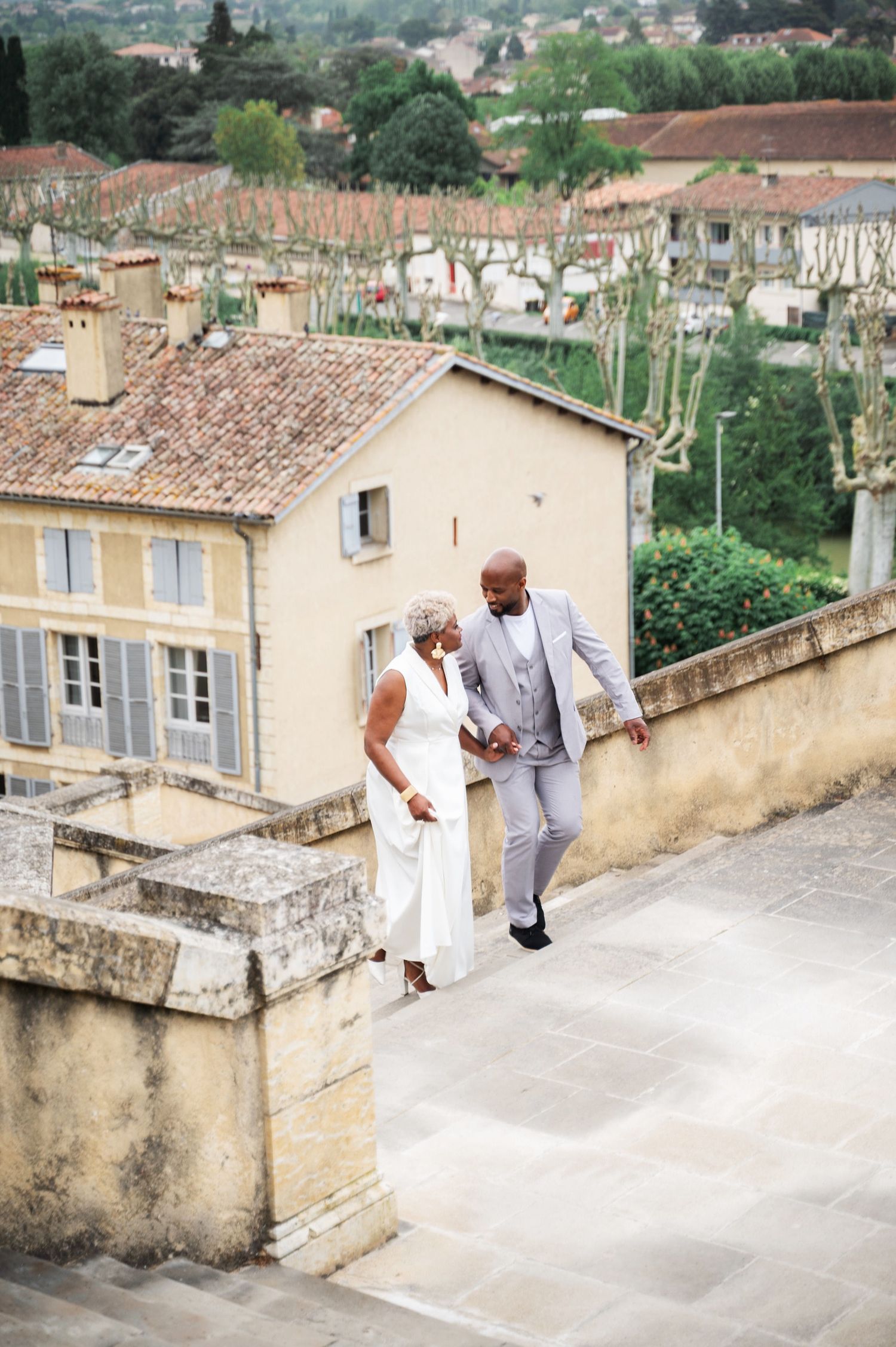 Black couple conversing on terrace overlooking French village, woman in white dress, man in gray suit.