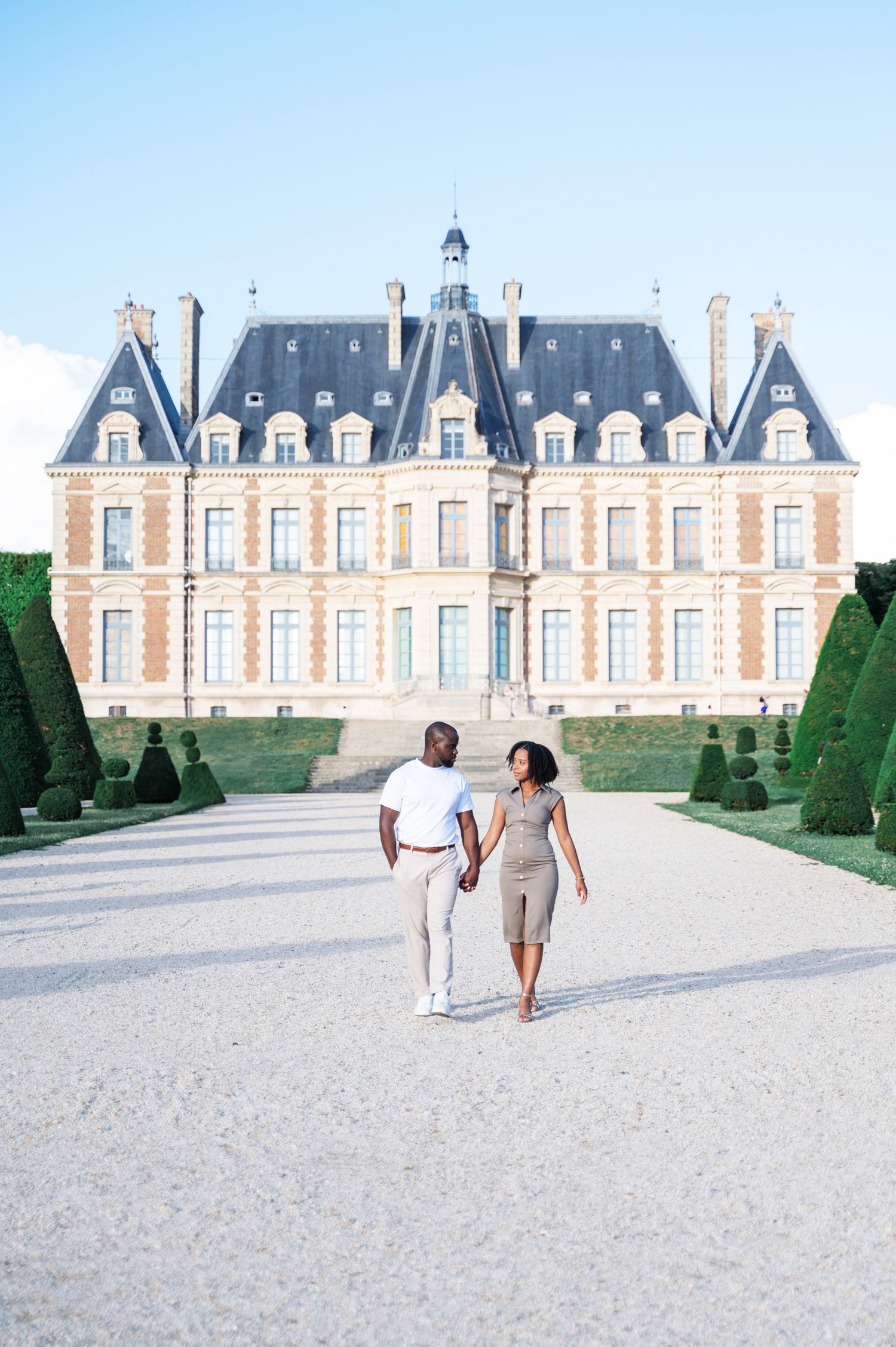 A couple holds hands walking toward the camera with a grand French château behind them on a sunny day.