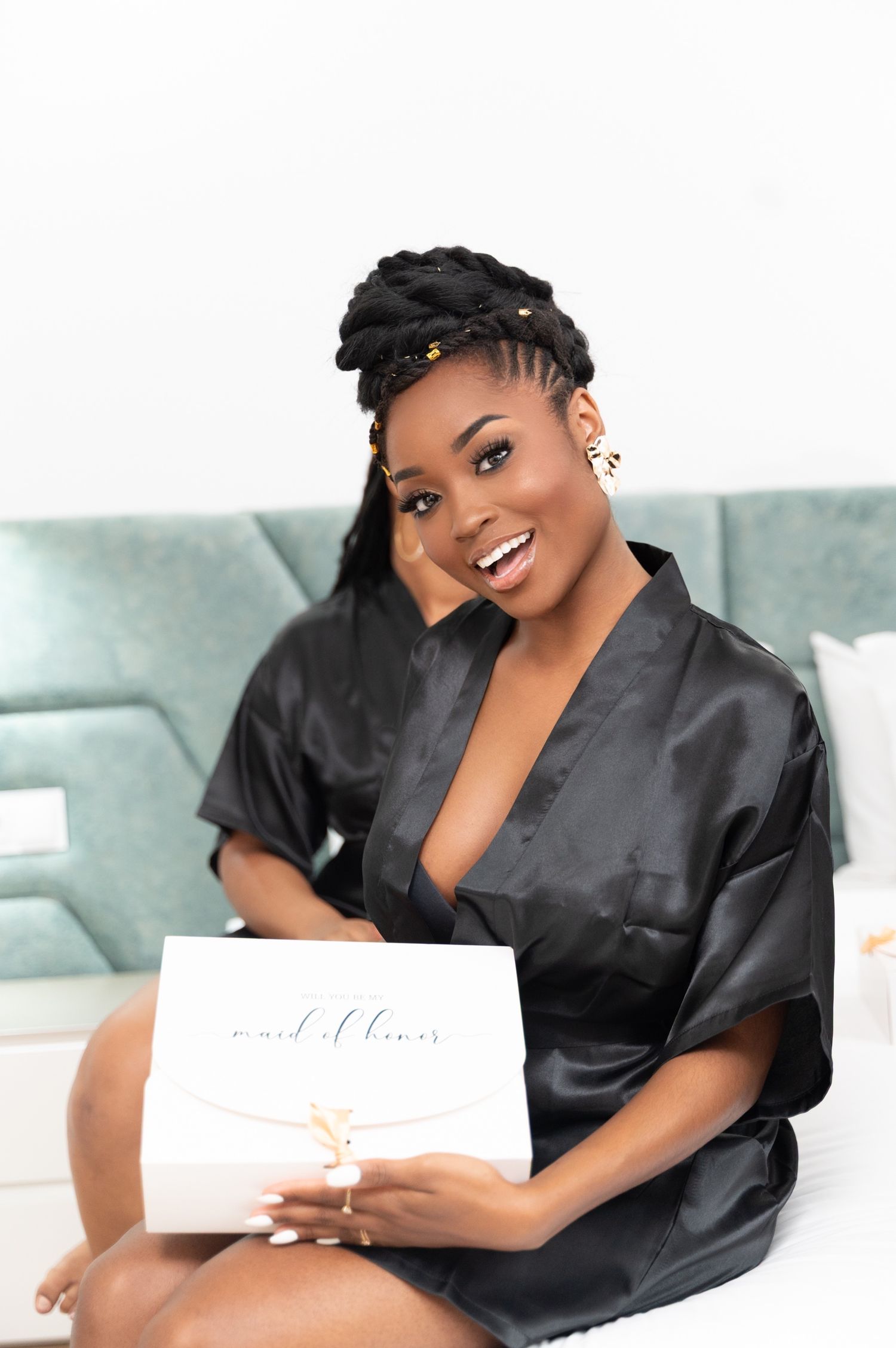 Happy woman in black satin robe holding a maid of honor gift box while sitting on a bed.