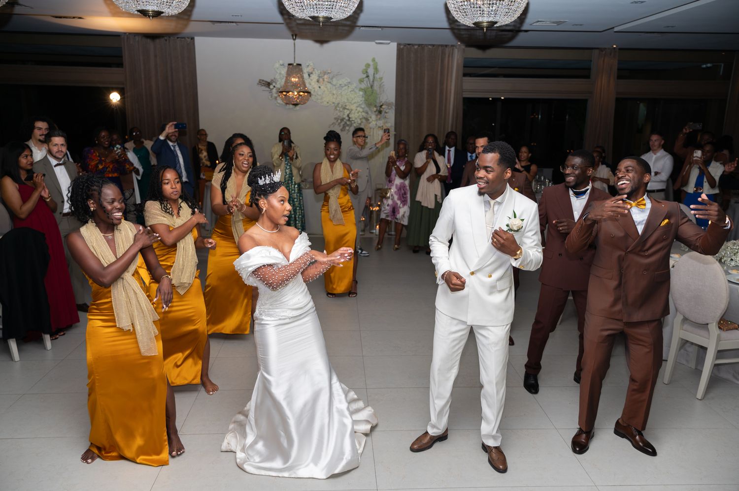 Bride and groom share first dance at wedding reception with bridesmaids in gold dresses and groomsmen watching.