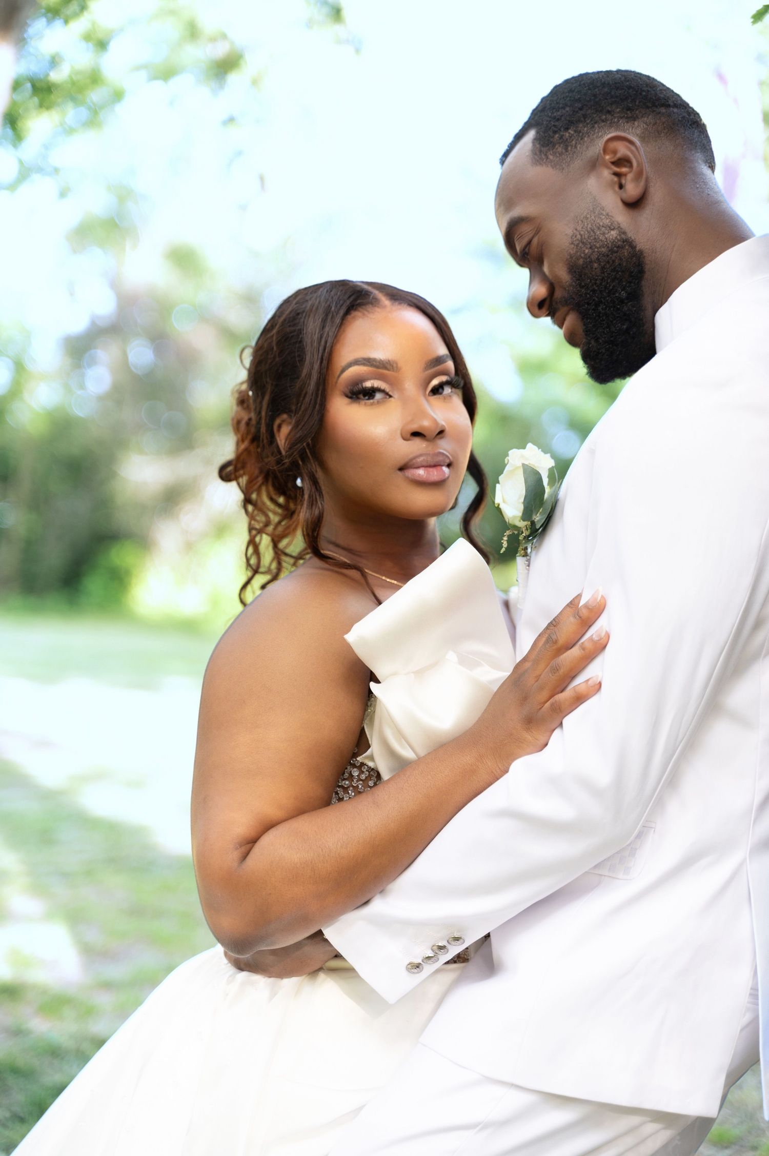 Radiant bride in white off-shoulder dress embracing her groom in a white suit in a sunlit garden.