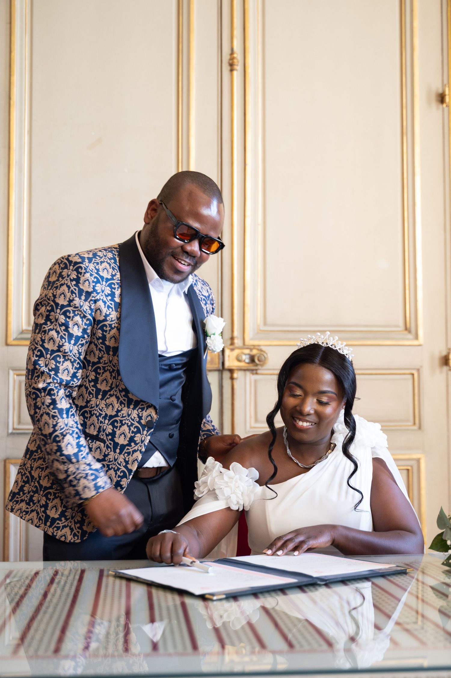 Smiling bride and groom sign wedding documents at a civil ceremony in an ornate gold-paneled room.