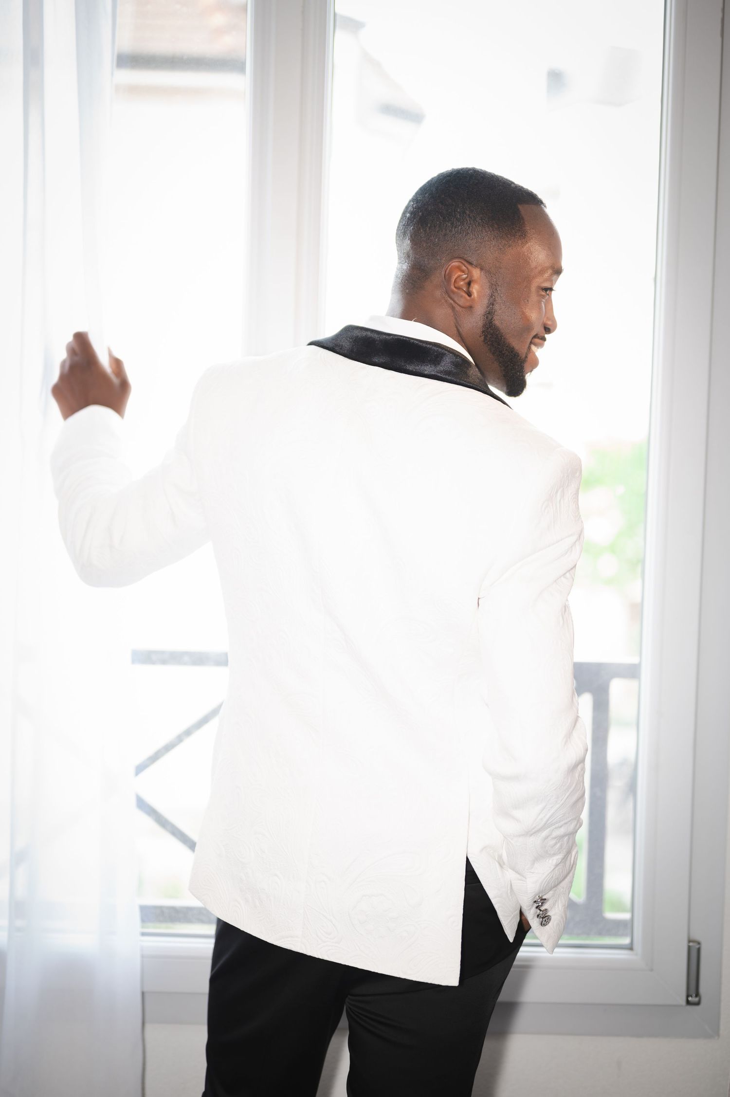 Groom in white tuxedo jacket with black collar looking out a bright curtained window smiling.