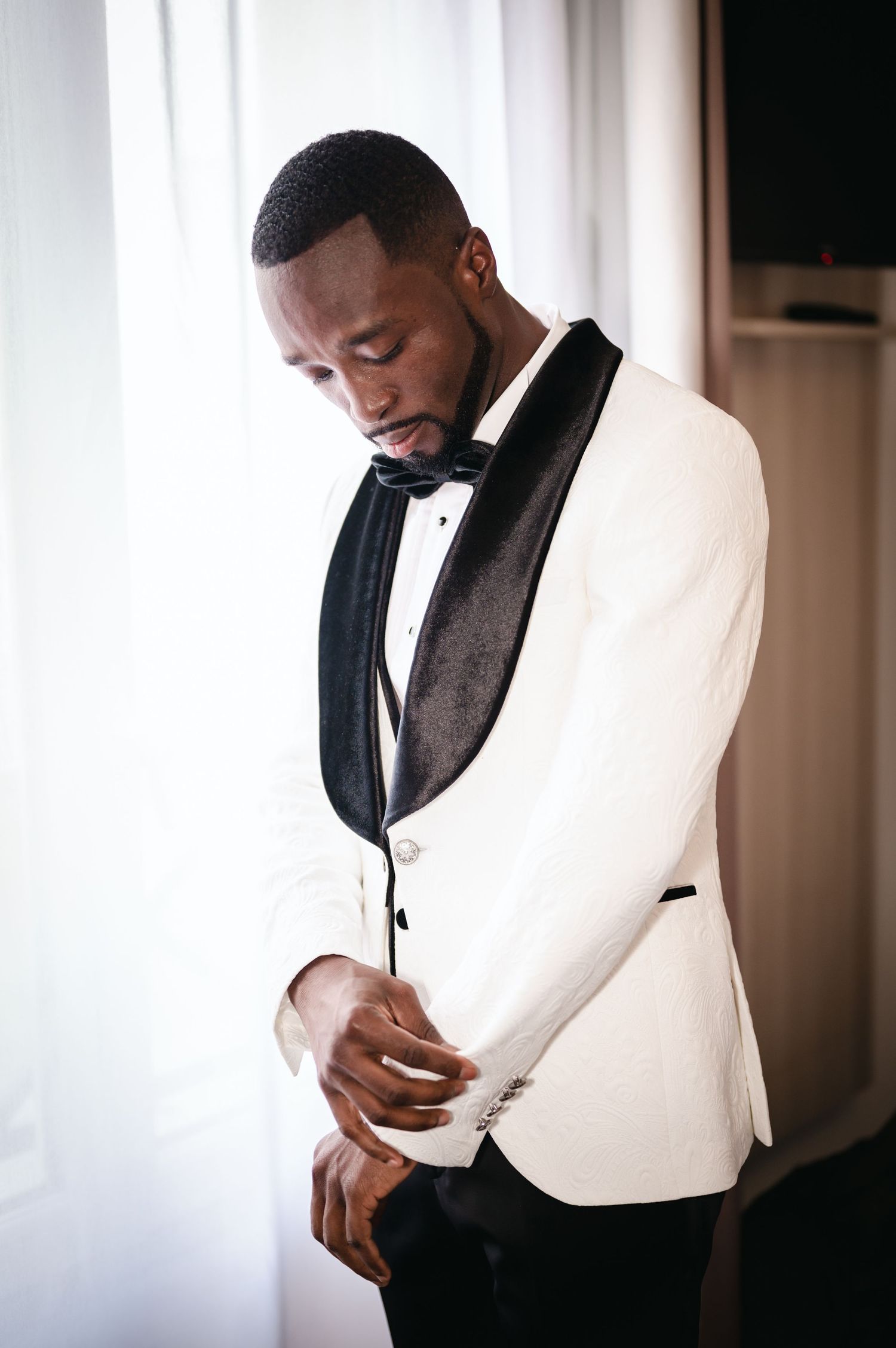 Groom in white tuxedo jacket with black lapels adjusting his suit near a bright window.