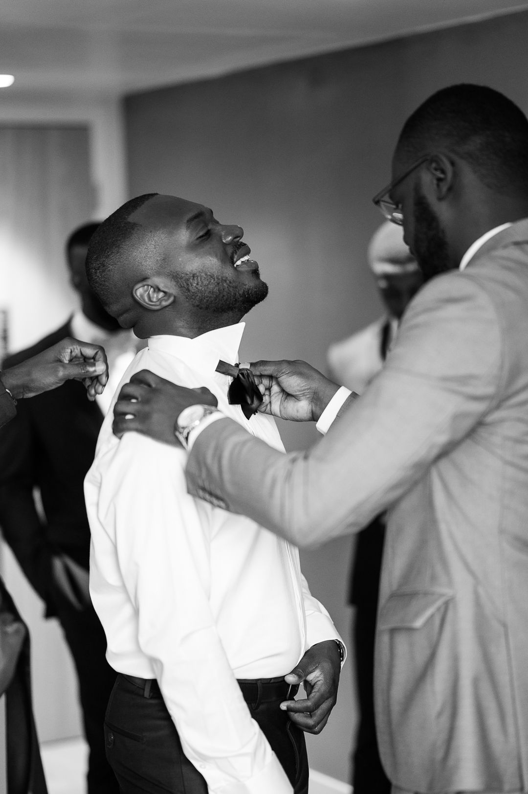 Groomsmen helping groom with bow tie while laughing in black and white wedding photo.