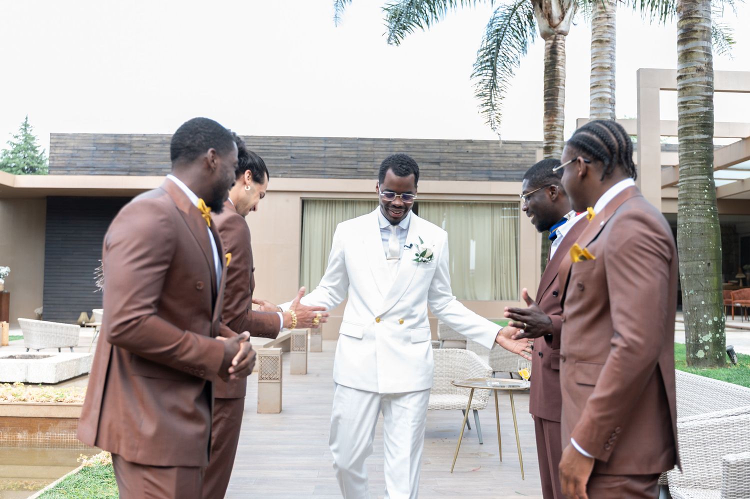 A groom in a white suit celebrates with groomsmen in brown suits with yellow accents outdoors at a wedding.