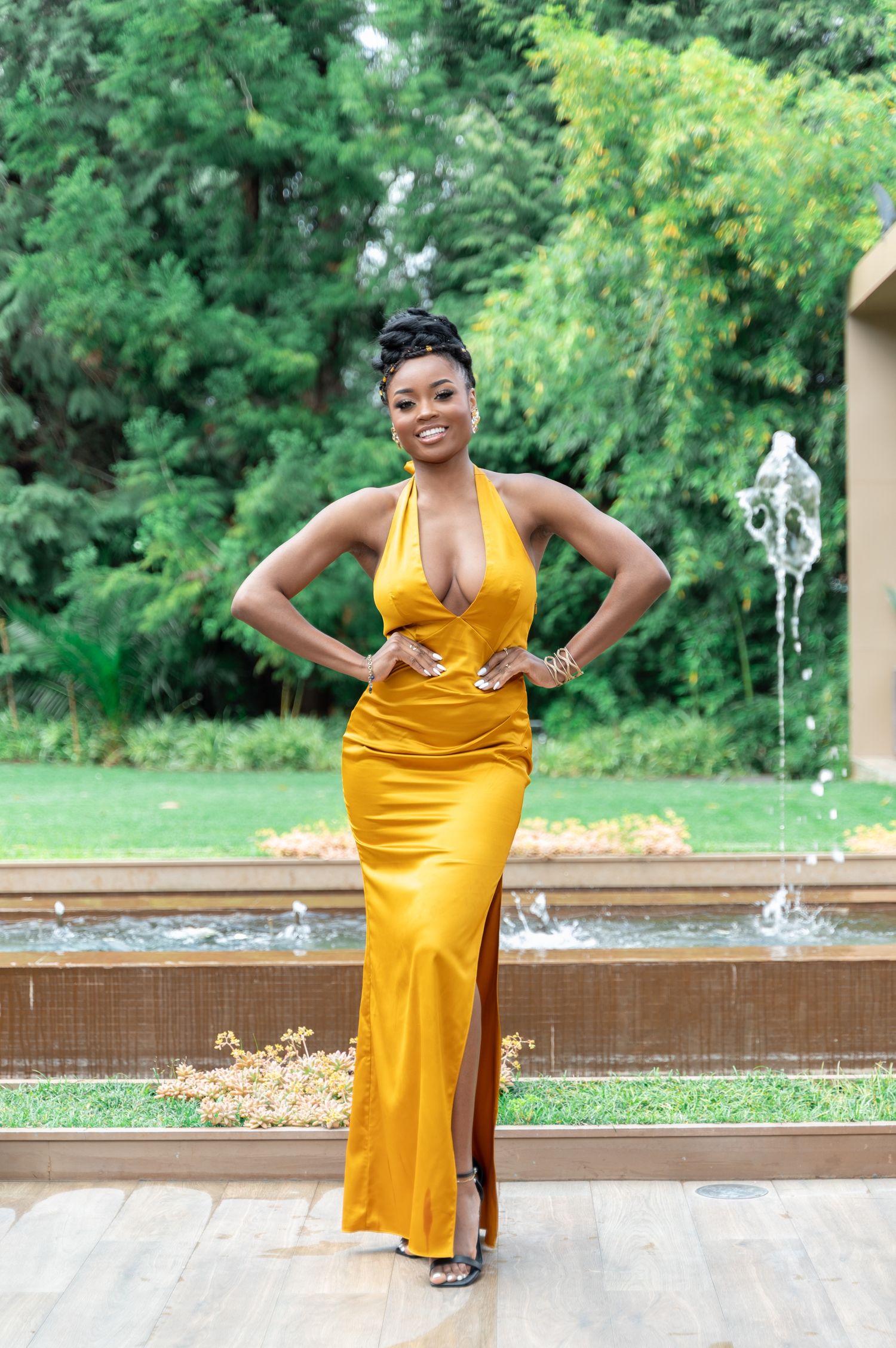 Woman in elegant gold halter maxi dress poses confidently by fountain in lush garden setting.