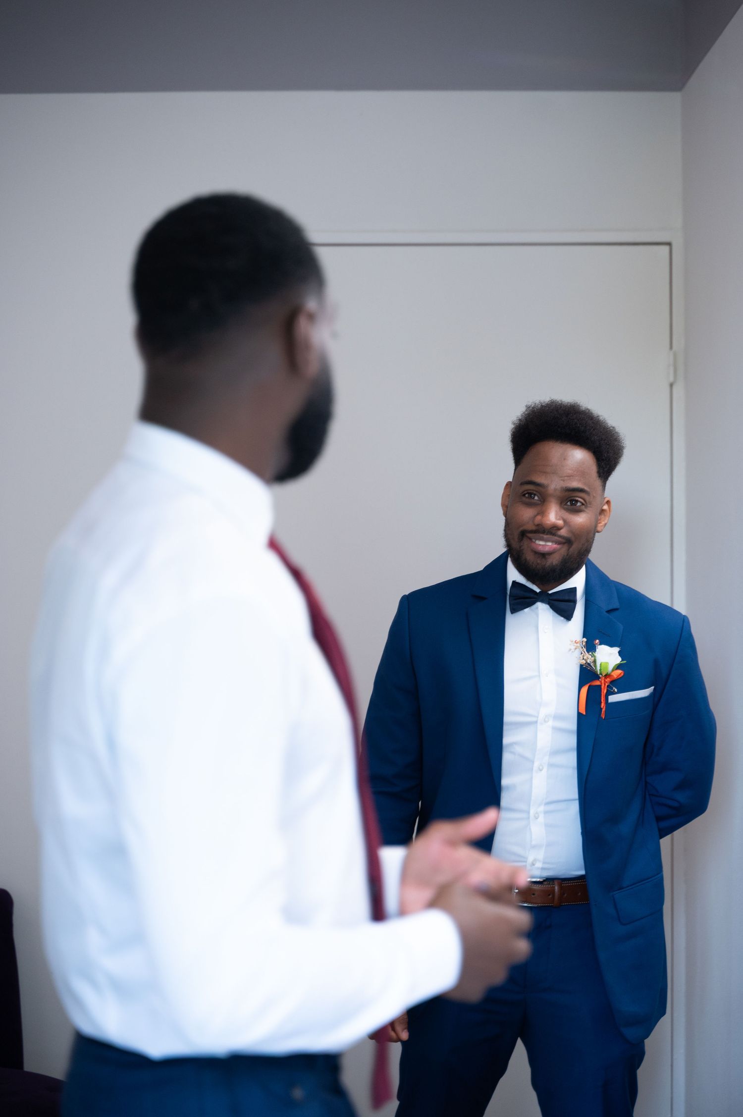 Black groomsman in navy suit with boutonniere smiling at another man in white shirt and suspenders in a room.