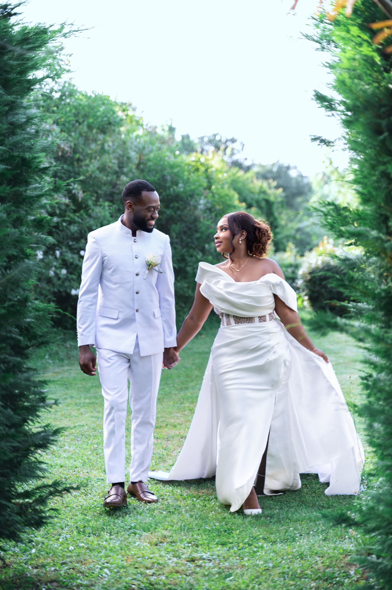 A Black bride and groom in matching white outfits walk hand in hand through a lush garden pathway.