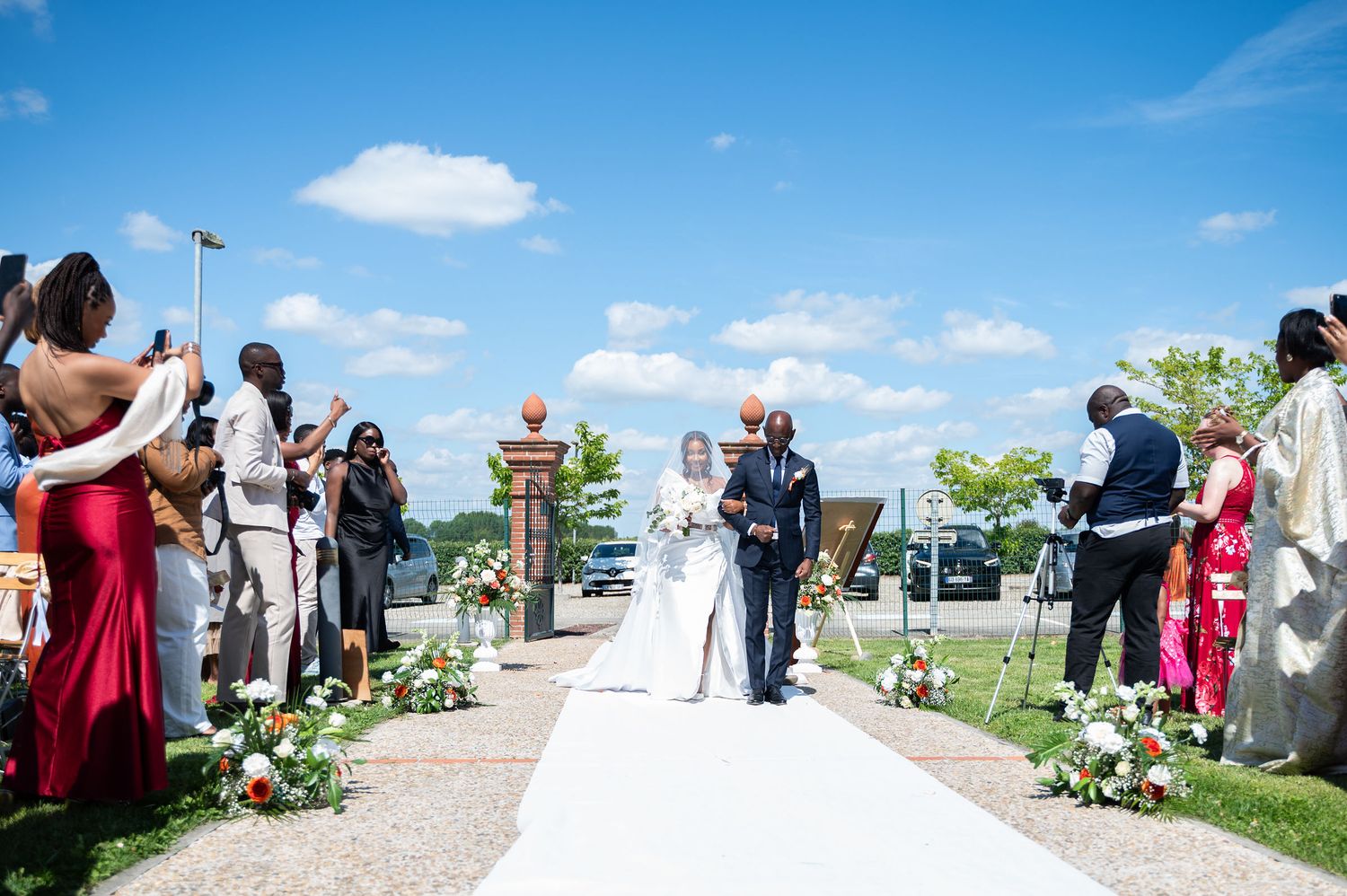 Bride in white gown walks down outdoor aisle with escort on sunny blue-sky wedding day.