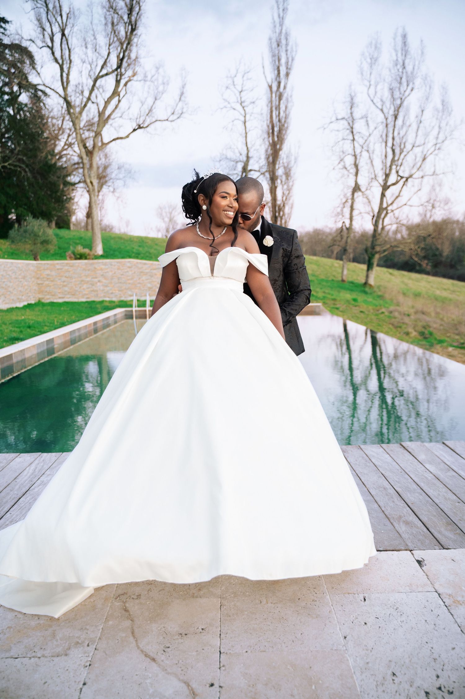 Bride and groom embracing by reflective pool in stunning white ball gown.