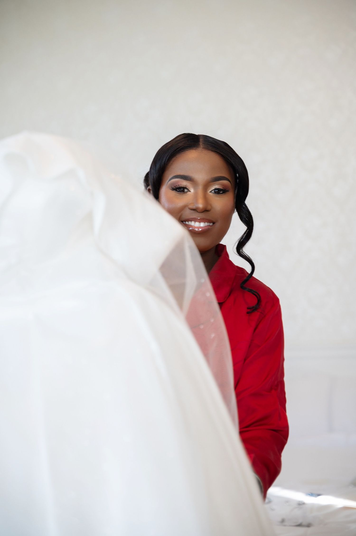 A smiling Black bridesmaid in a red jacket peeks through a white wedding veil during bridal preparations.