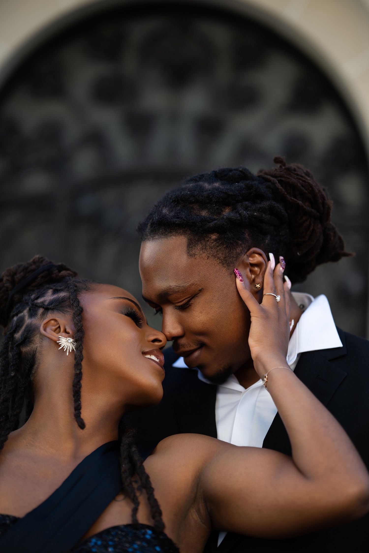 A romantic couple in formal attire sharing an intimate moment, foreheads touching, with dreadlocks and elegant accessories.