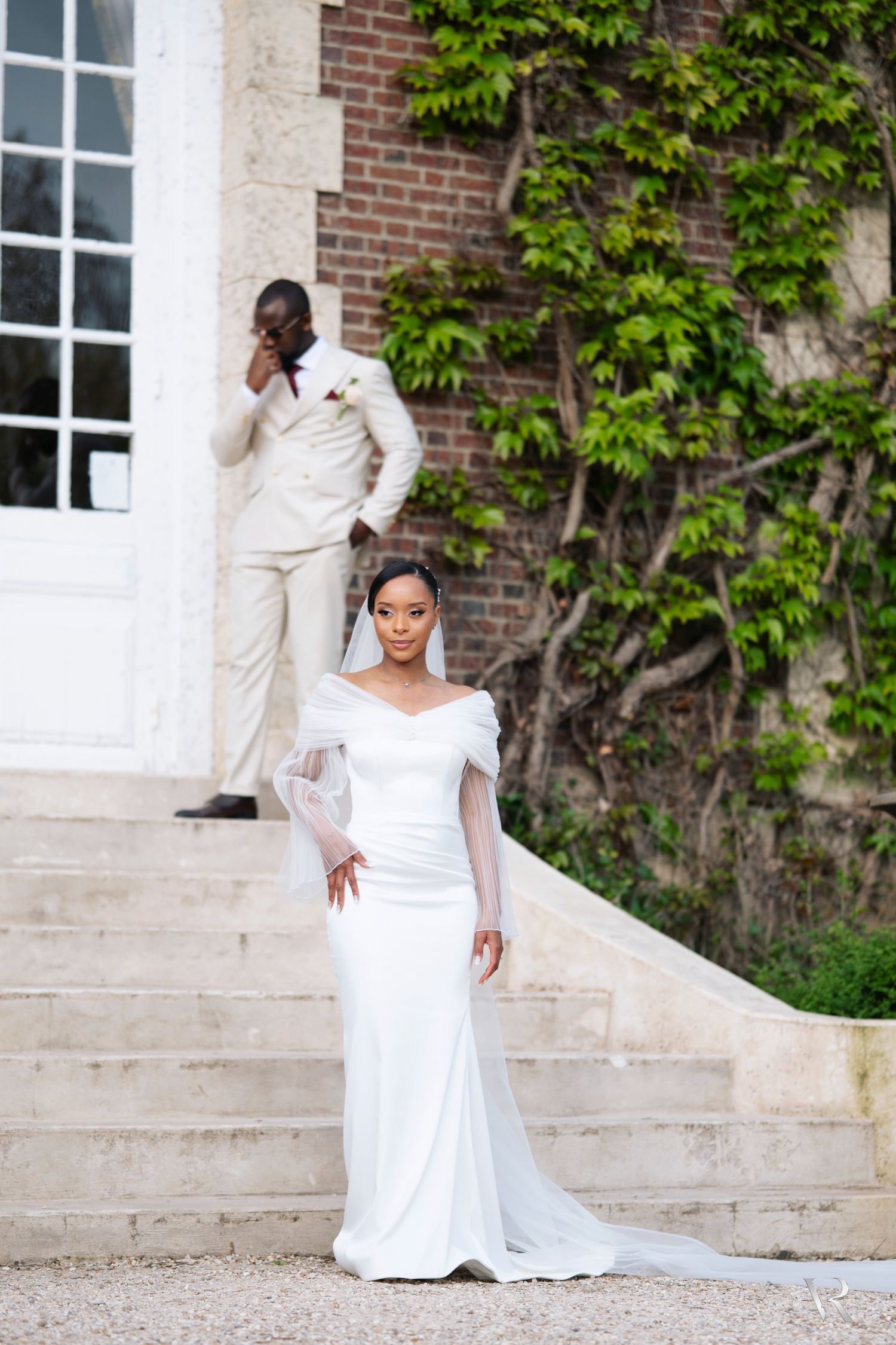 Bride in elegant white off-shoulder gown with veil stands on steps as groom in cream suit poses behind her.