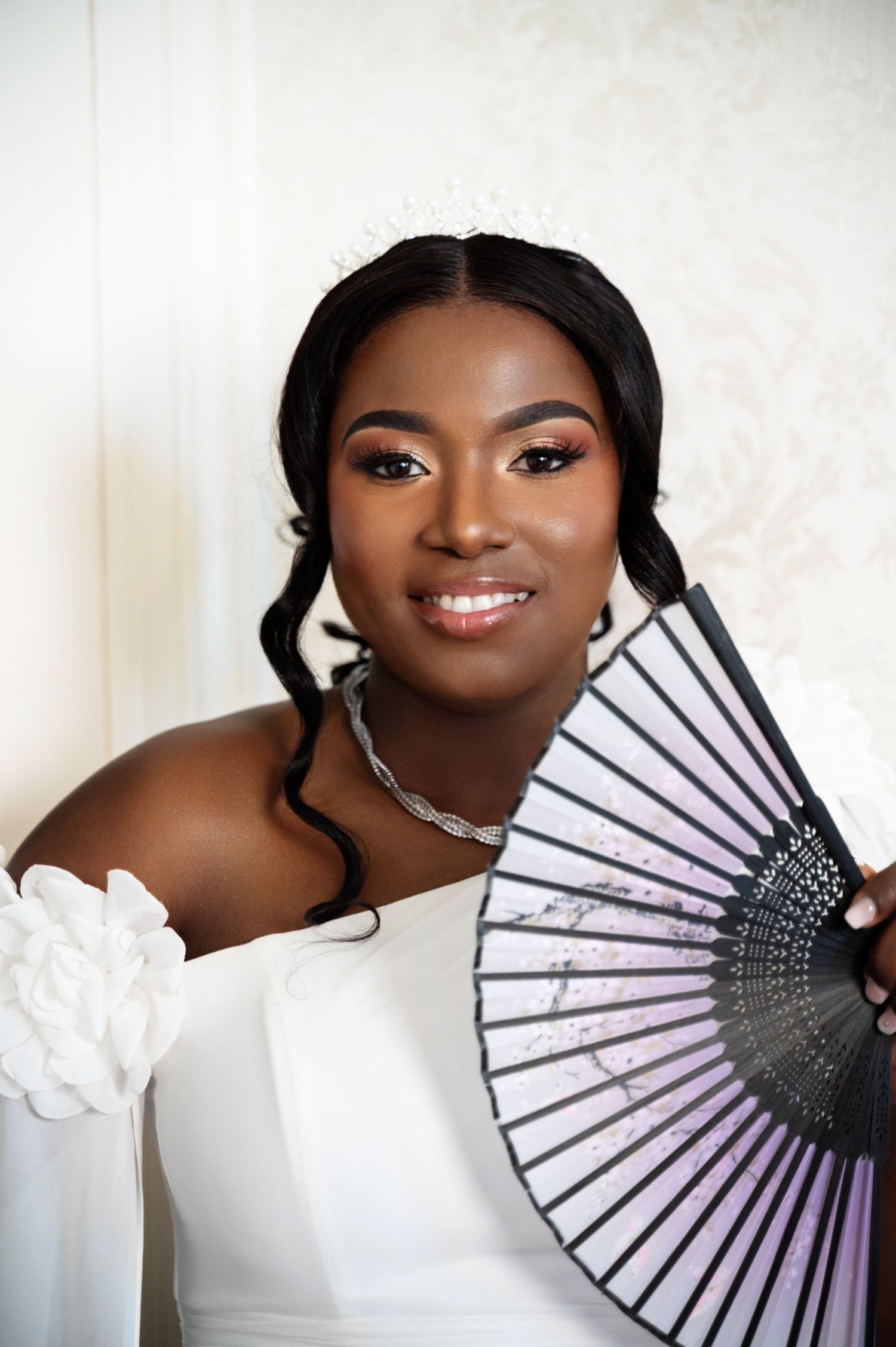 An elegant Black bride in a white off-shoulder gown holds a decorative fan, wearing a tiara and silver necklace.
