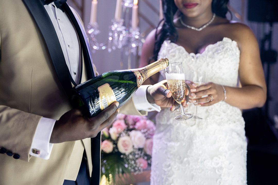 Groom pouring champagne into glasses held by the bride in a white lace wedding gown at reception.