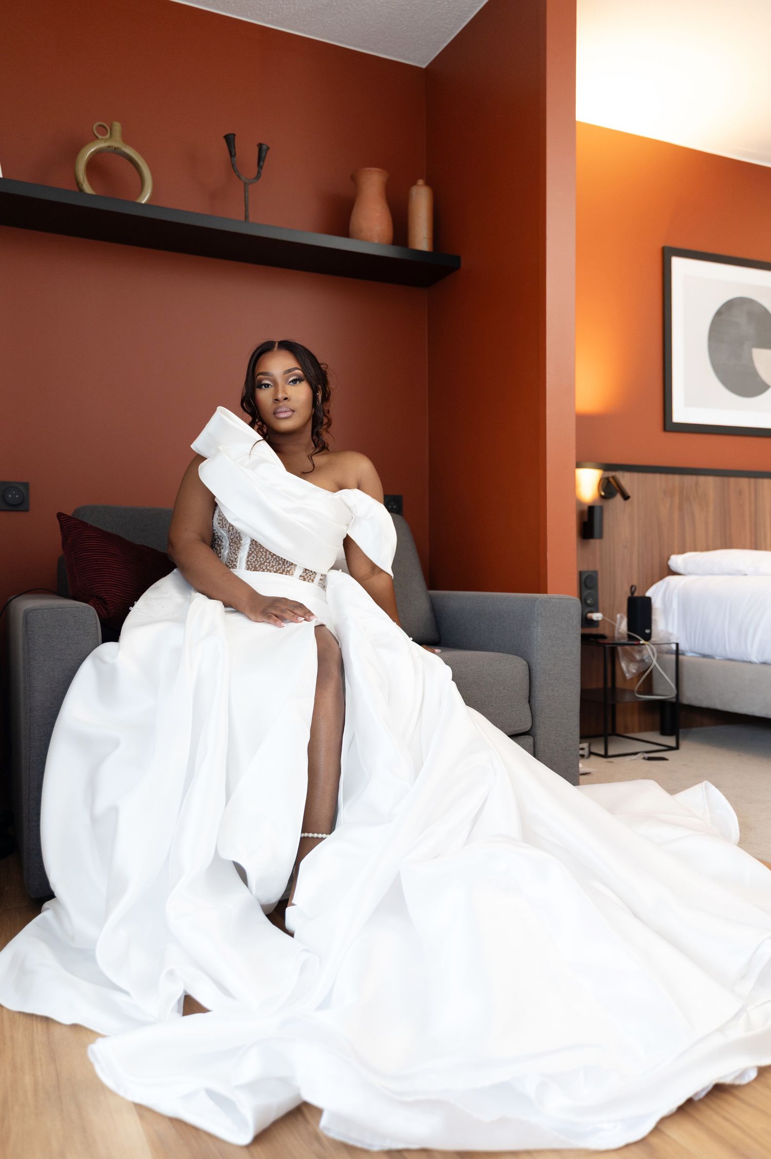 Glamorous Black bride in dramatic off-shoulder white gown with train poses in warm terracotta-toned hotel suite.