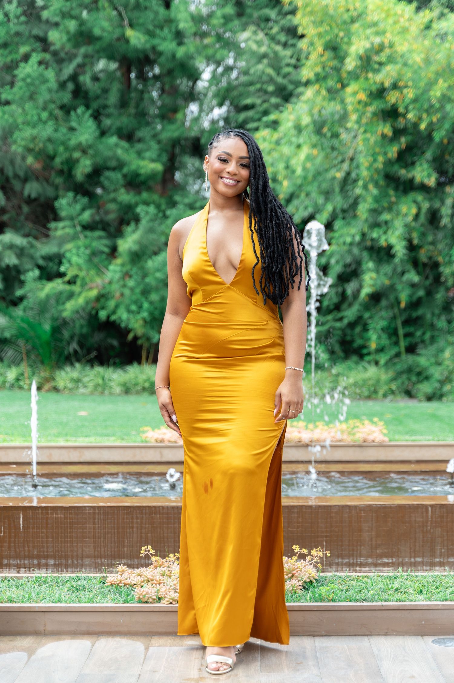 Woman in golden yellow halter maxi dress posing by fountain in lush garden.