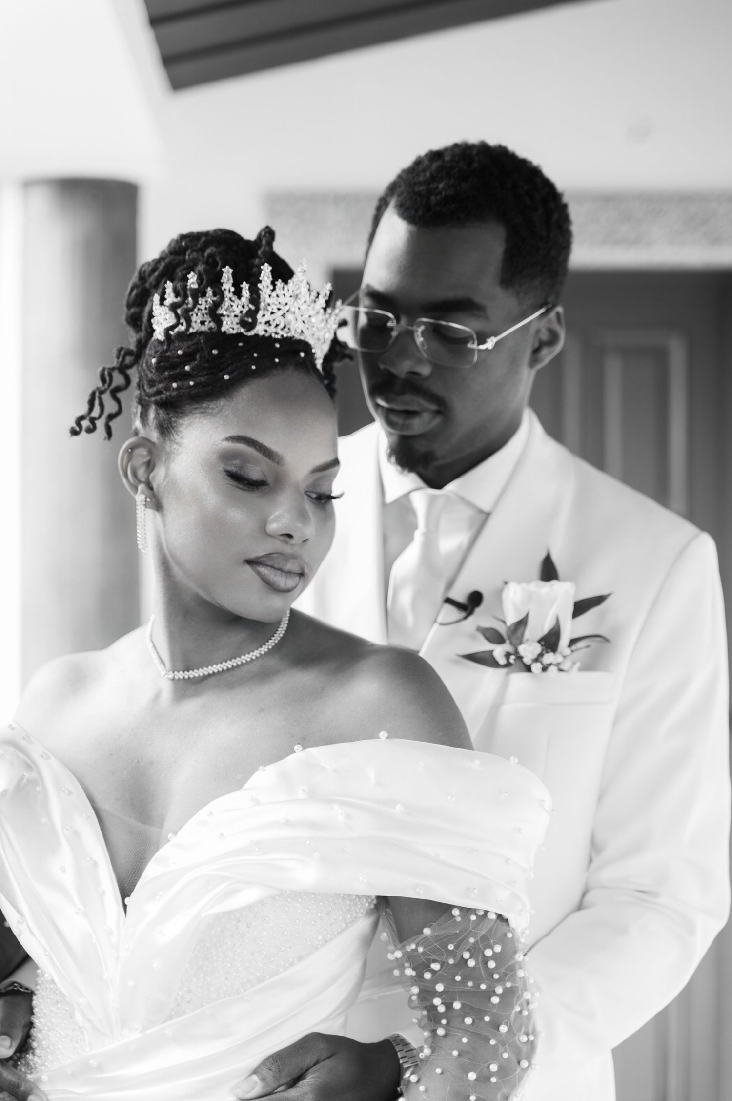 Black and white portrait of bride in pearl gown and crown with groom embracing her from behind.