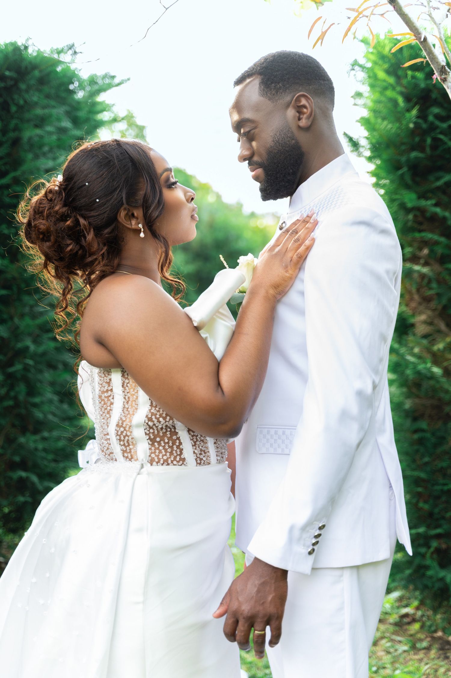 Black bride and groom in white wedding attire sharing an intimate romantic moment in a lush green garden.