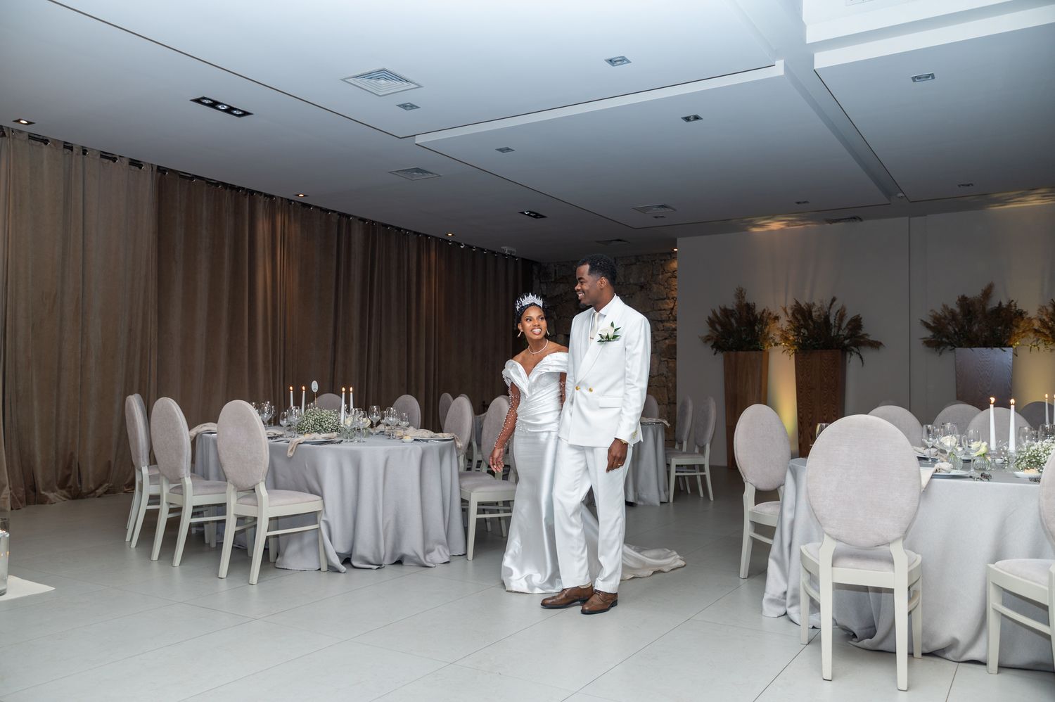 Bride and groom enter elegant white wedding reception hall with round tables and ghost chairs.