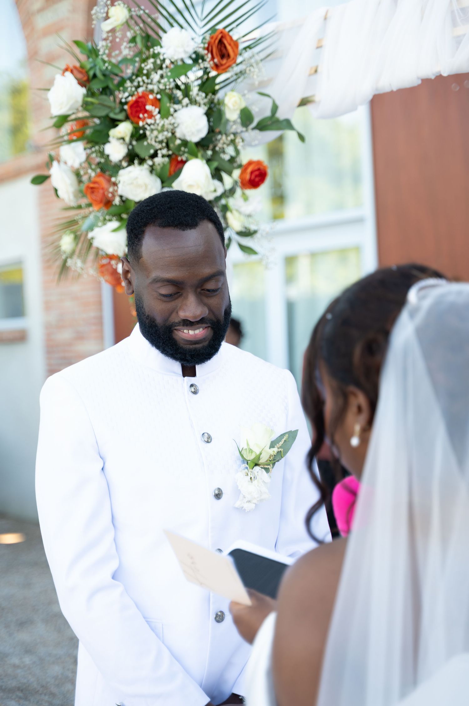 Smiling groom in white suit listens to bride recite vows under floral arch.