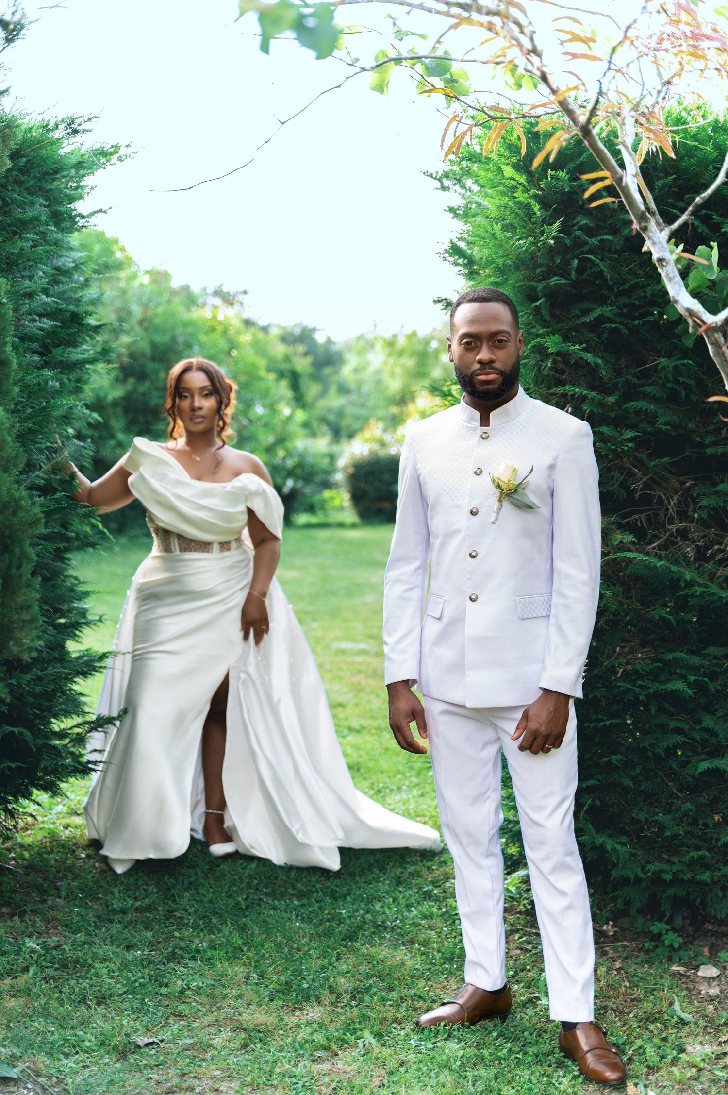 A Black bride and groom pose separately in a lush garden, both wearing elegant white wedding attire.