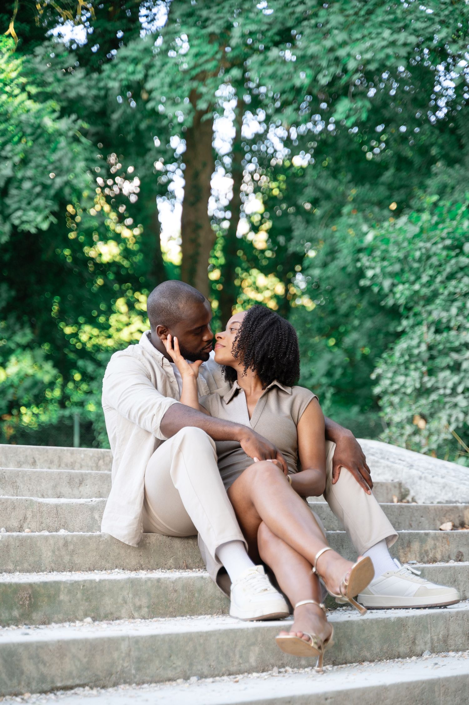 A romantic Black couple sits closely together on stone steps surrounded by lush green trees in a park.