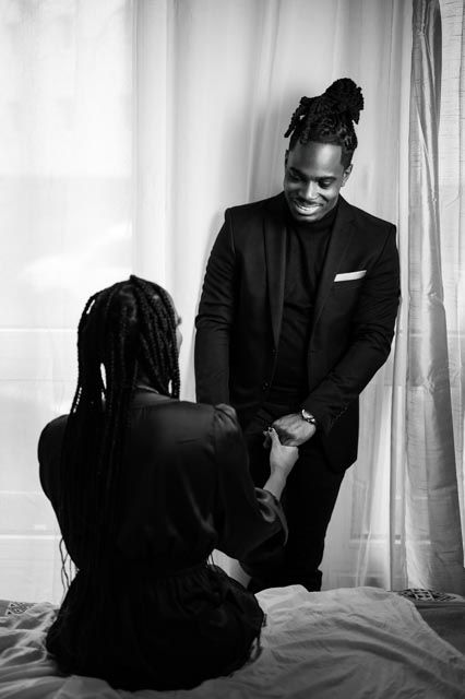 Couple in elegant black attire share intimate moment, man in suit holds seated womans hand.