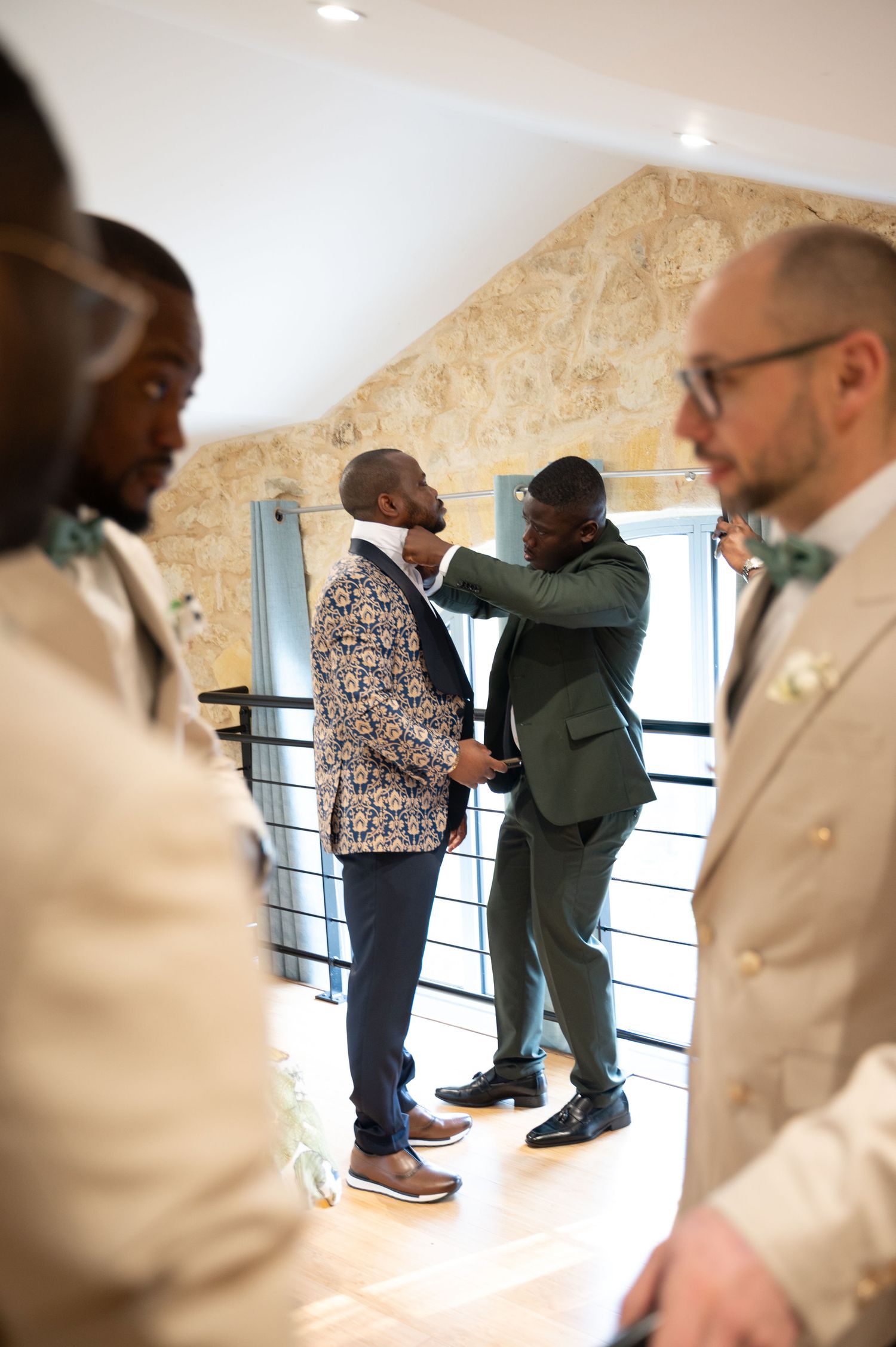 A groom in a floral blazer gets his boutonniere adjusted by a groomsman in a green suit before the wedding ceremony.
