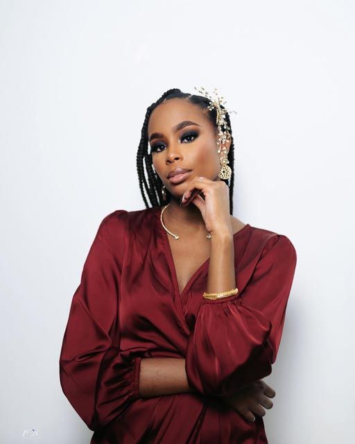 Young woman in burgundy satin dress with box braids and gold accessories poses confidently against white background.