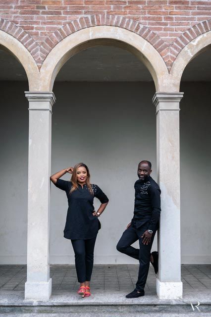Two stylish people in black outfits pose under elegant stone arches with brick accents in a classic architectural setting.