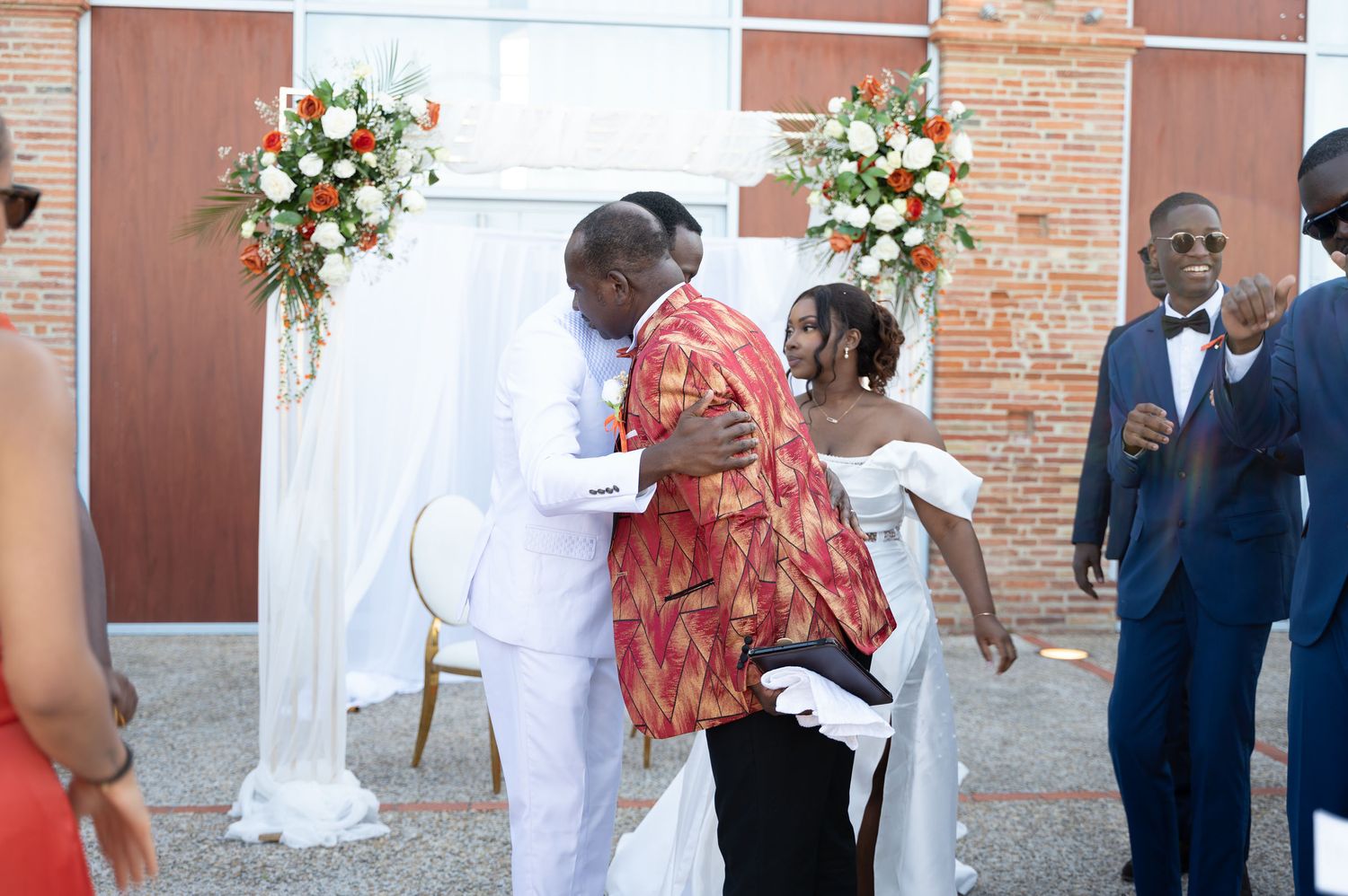 Groom in white suit embracing a guest in colorful African print attire at outdoor wedding ceremony.