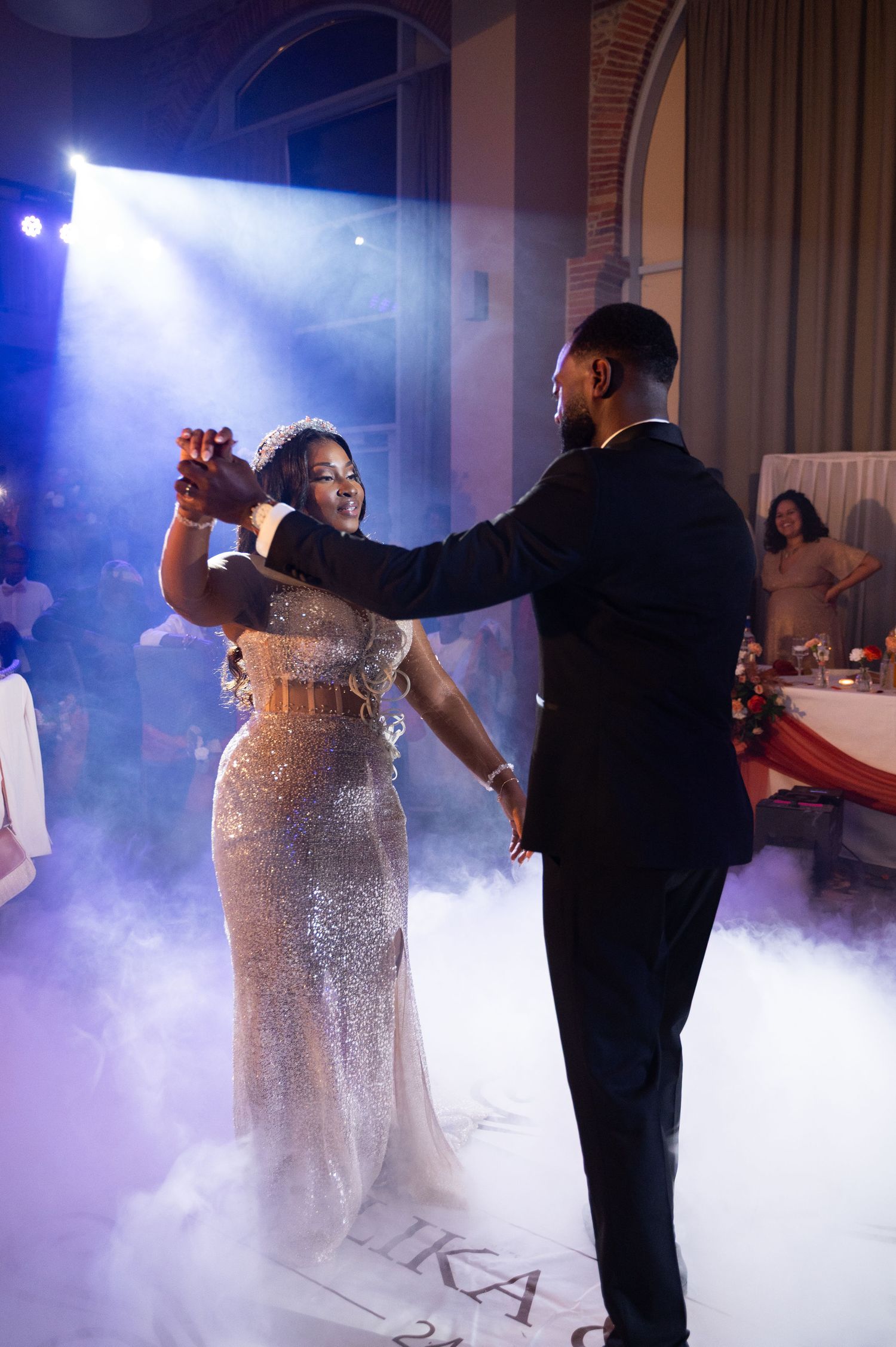 Bride in sparkling sequin gown and groom in black tuxedo share romantic first dance surrounded by atmospheric fog.