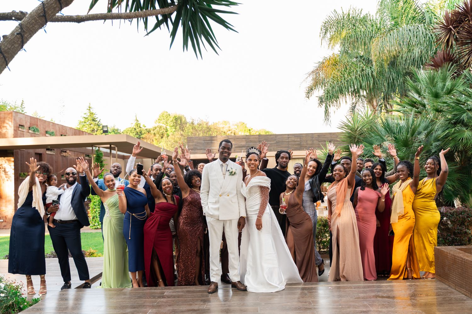 Bride and groom celebrate with large wedding party outdoors, bridesmaids in colorful gowns.