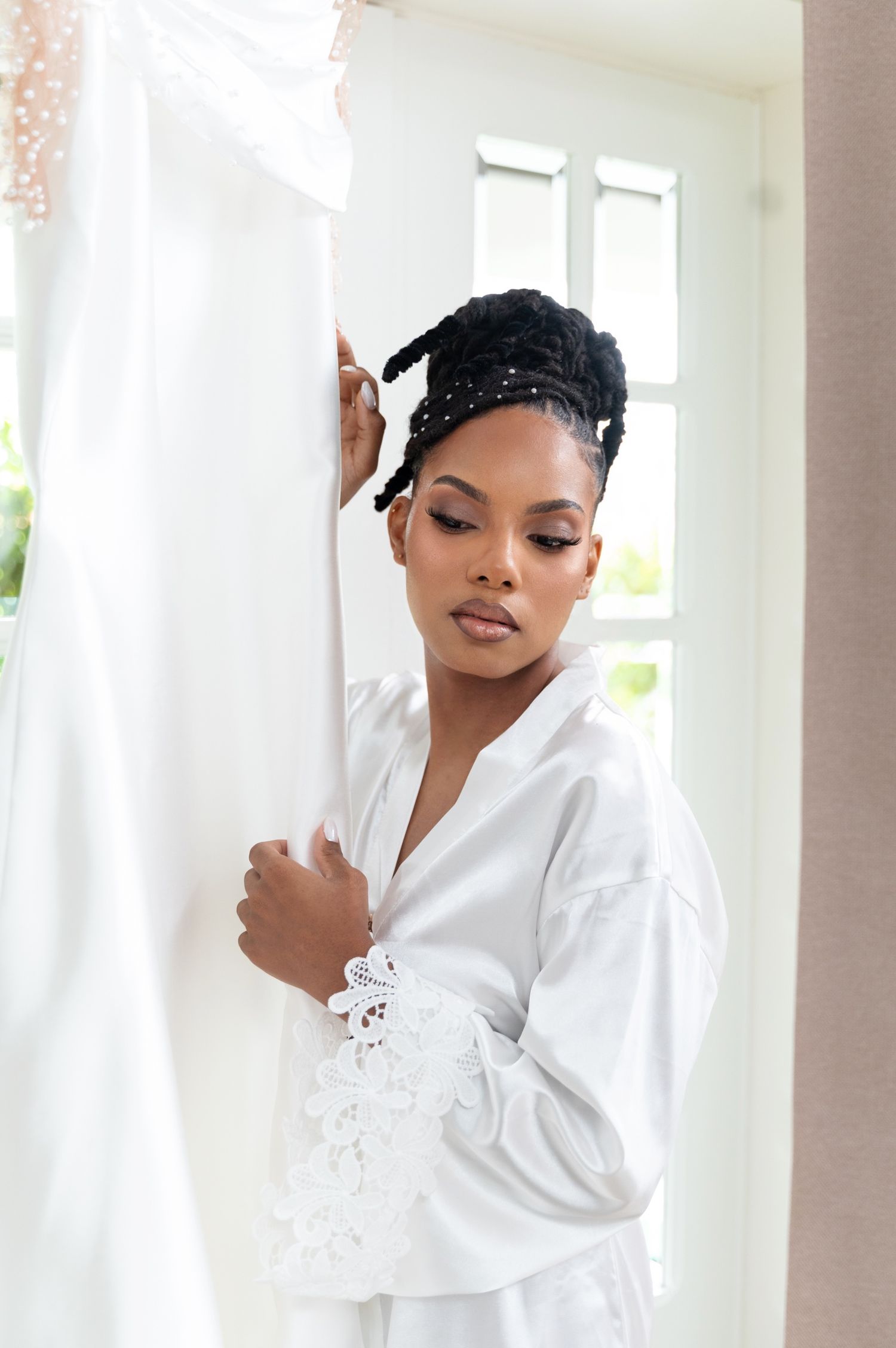 Beautiful bride in white satin robe gazing at her wedding dress hanging nearby.