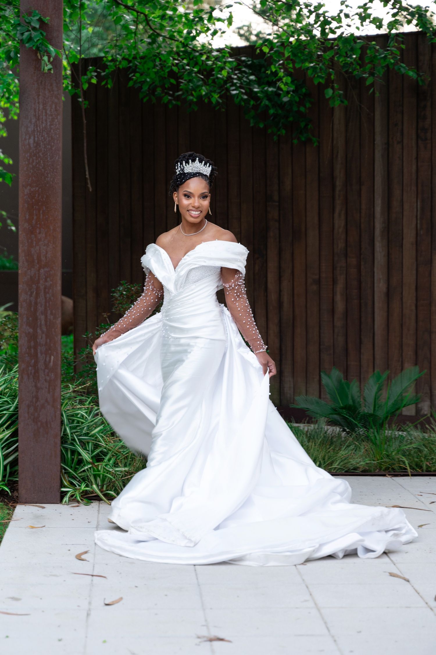 Bride in elegant off-shoulder white gown with pearl sleeves and crystal crown poses outdoors.