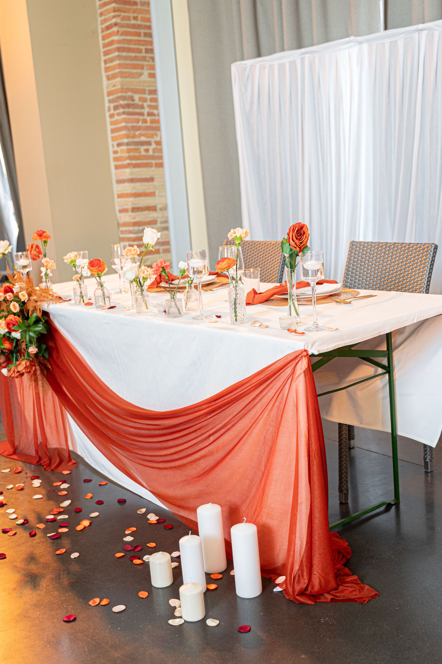 Wedding sweetheart table with orange draped fabric, roses, candles, and rose petals on dark floor.